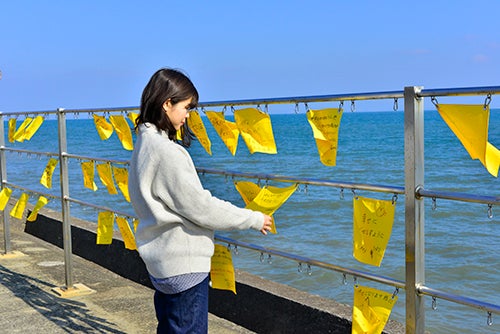 海沿いの駅のホームに掲げられたたくさんの黄色いハンカチを見る女性