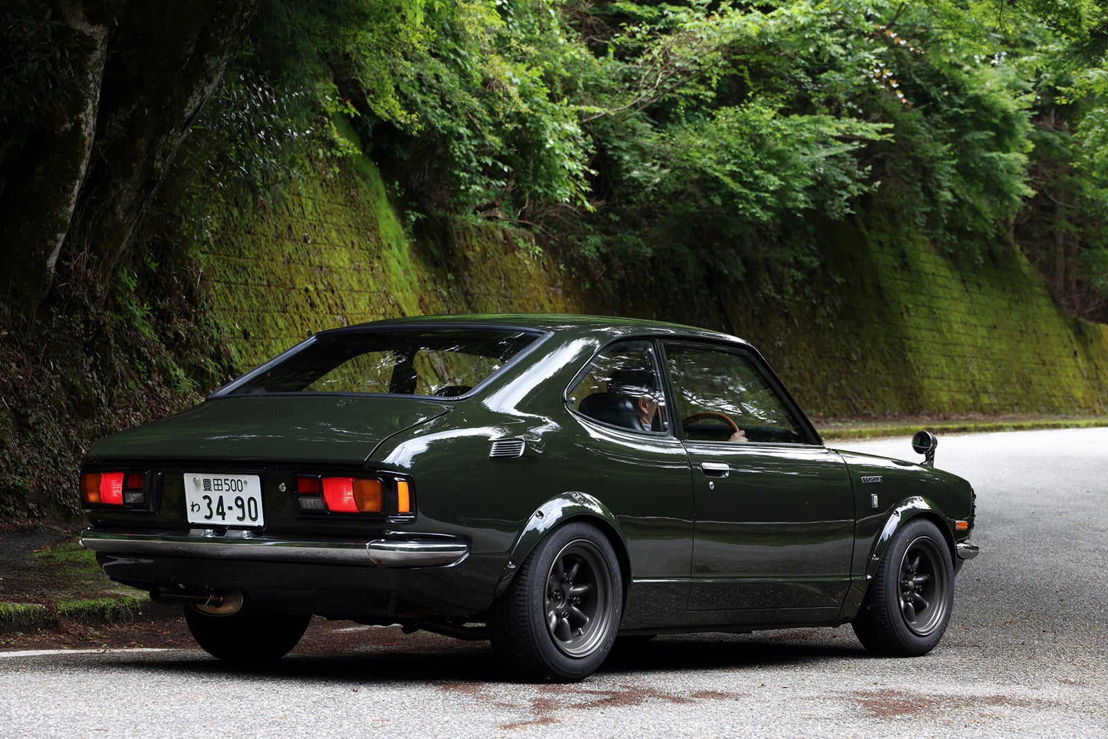 緑に囲まれた道路に停車する1974年式トヨタ・カローラレビンTE27型モスグリーンの後方ビュー。Vintage club by KINTOのクラシックカー