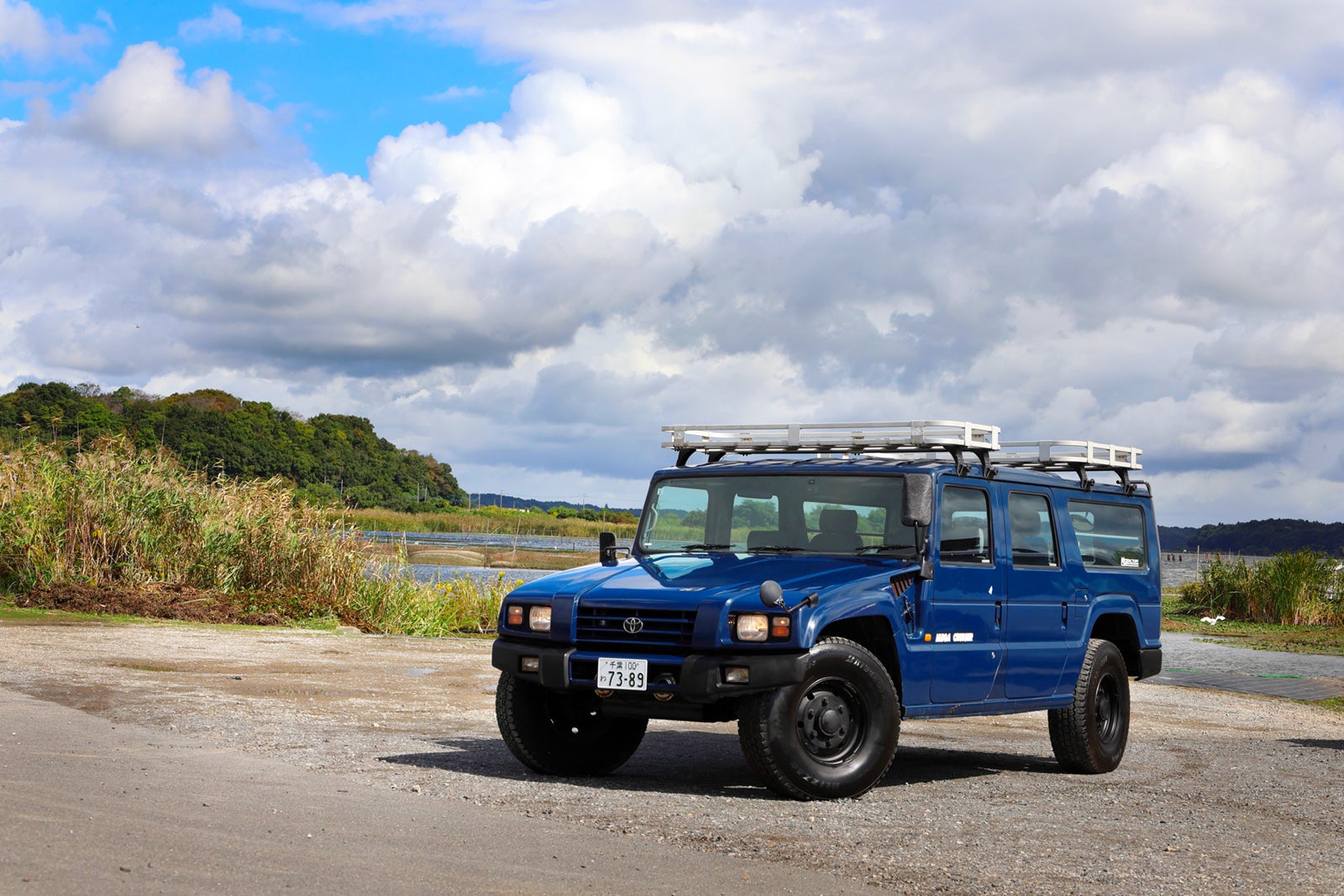 印旛沼のほとりに佇むブルーのトヨタ・メガクルーザー。左前3/4外観