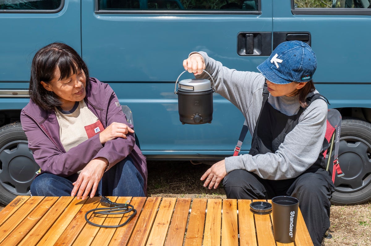クルマの前でイスに座り電気調理器を眺める女性2人