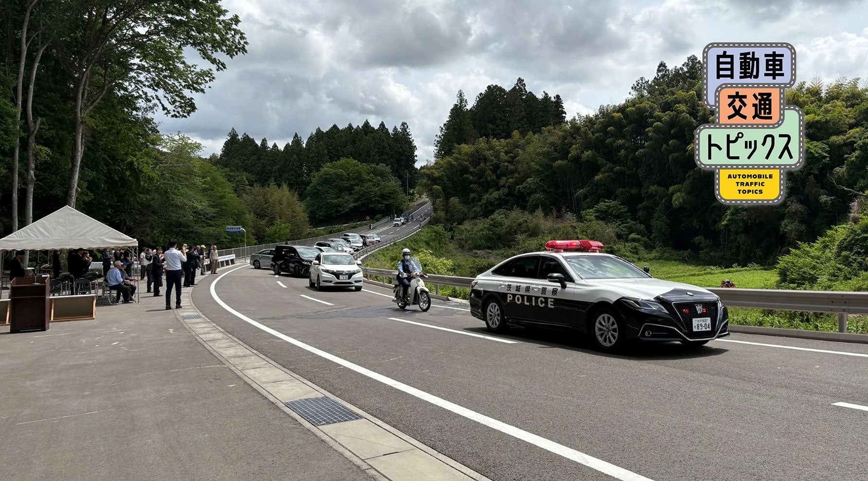 開通したバイパスで実施された通り初めの様子。パトカーを先頭に、バイク・乗用車などが新しい2車線道路を走行し、左手のテント付近では関係者が見守っている