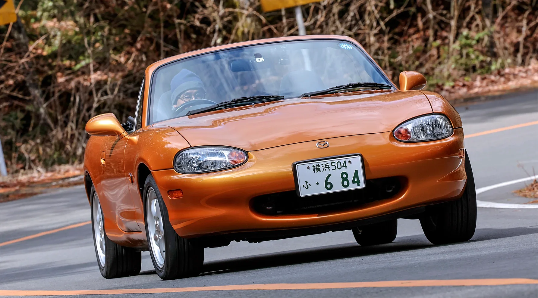 峠道の緩やかなカーブをオープン状態で走行する、エボリューションオレンジマイカのマツダ・ロードスター（NB型）。フロントをやや斜め前に向け、運転席にはニット帽を被った男性が座っている。背景には冬枯れの木々や傾斜地が見える