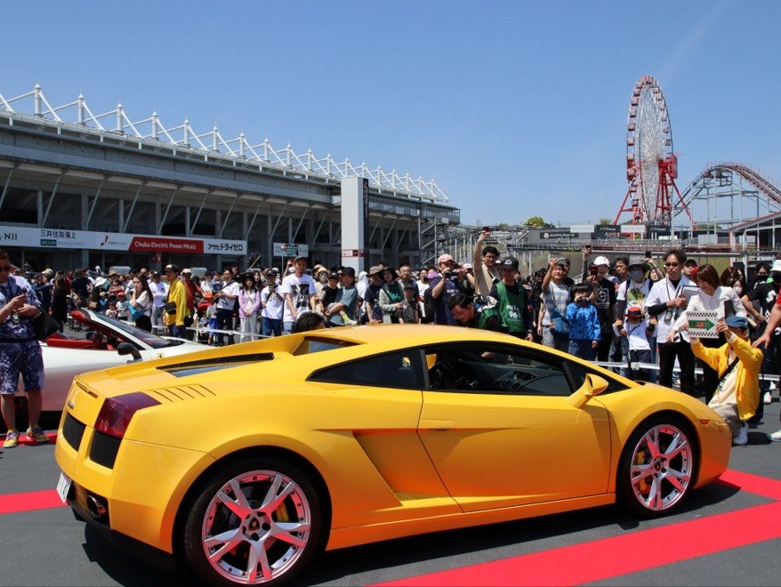 鈴鹿サーキットパークのGPスクエアで、グランドスタンドと観覧車を背景に展示される黄色いランボルギーニと、それを取り囲む来場者たち