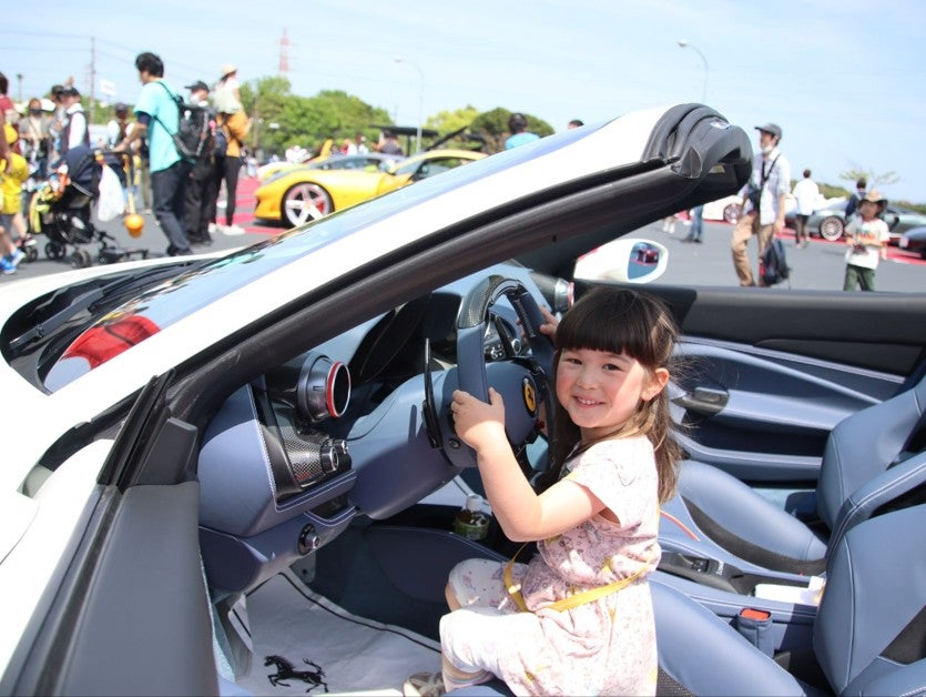 オープンカーのフェラーリの運転席でハンドルに手をかけ、カメラに向かって笑顔を見せる女の子。