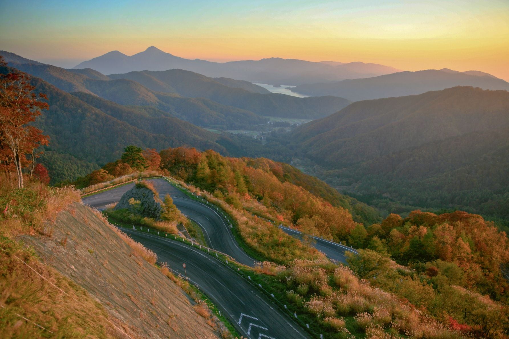西吾妻スカイバレー・東鉢七曲りの山岳道路。紅葉に包まれた急カーブの道が夕陽に照らされ、遠くに連なる山々と青空が広がる絶景ドライブコース