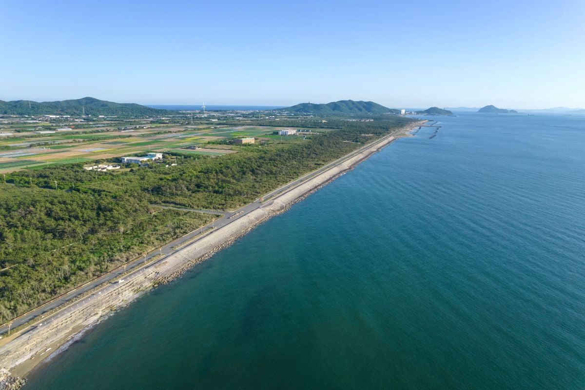 伊良湖岬から北上して西ノ浜へ向かう海岸地域の空撮写真。右側には澄んだ青い海、左側には緑の畑と森林、中央には海岸に沿って伸びる直線道路が見える