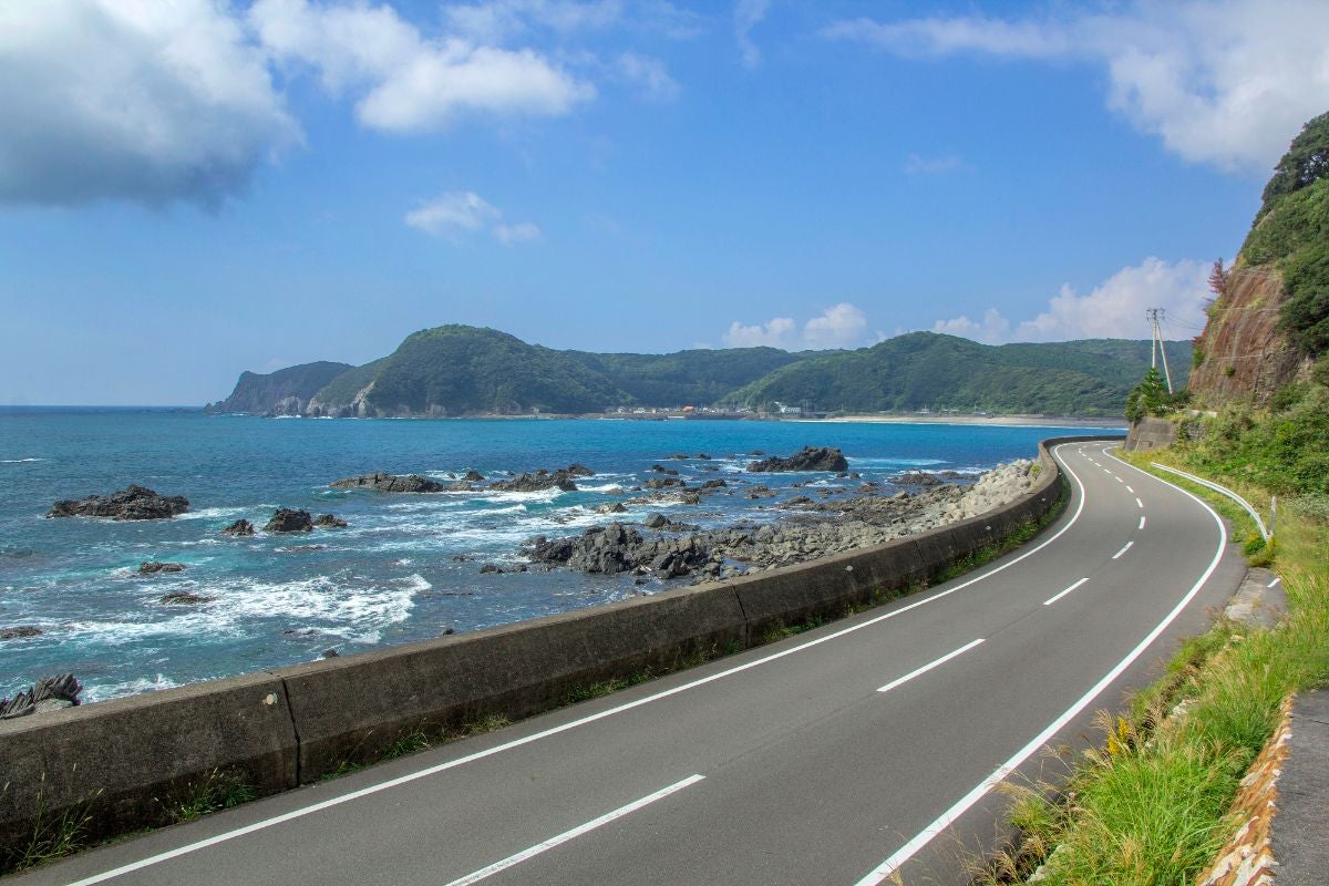 高知県・足摺サニーロードの海沿い道路。右側に緩やかなカーブを描く舗装路、左側には岩場と波打つ太平洋が見える