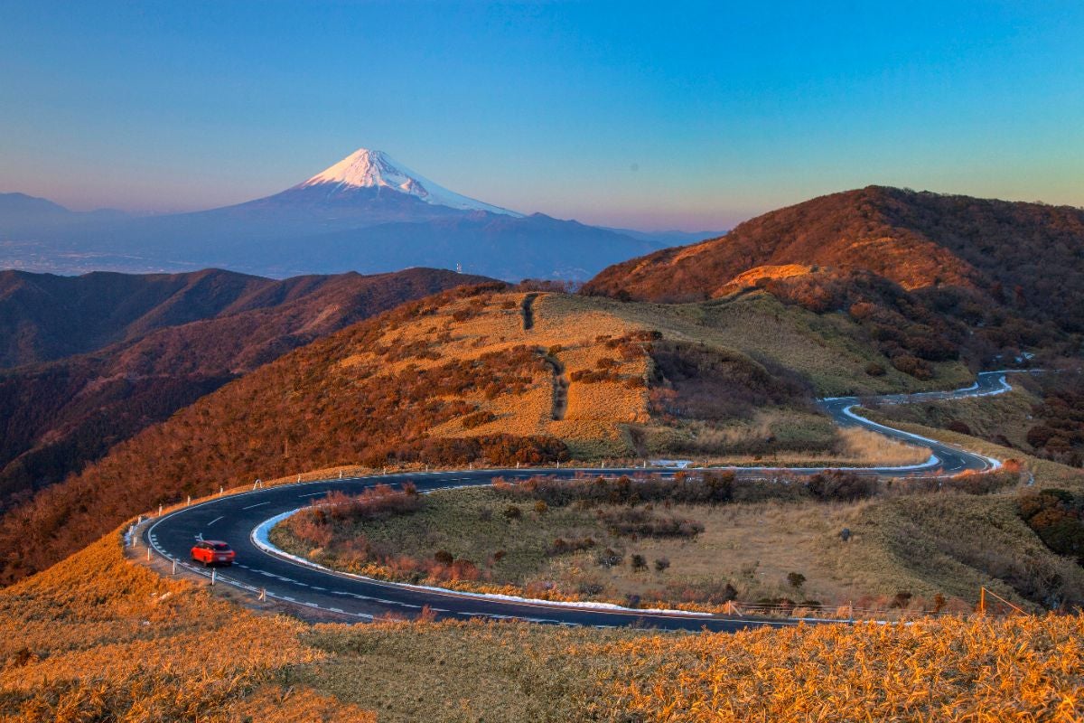 夕暮れの西伊豆スカイラインを走る赤い車と、黄金色に輝く稜線。その奥には雄大な富士山がそびえる
