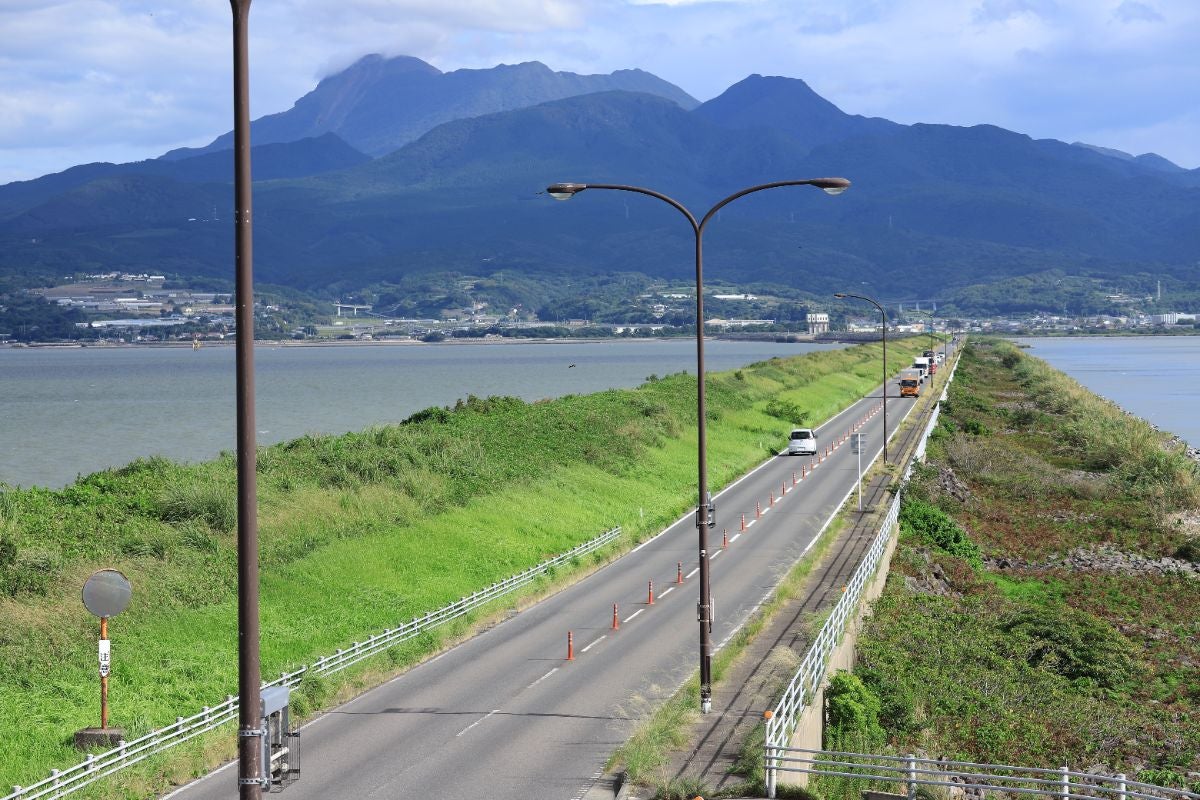 諫早湾干拓堤防道路の直線コース。道路の先には雄大な雲仙の山々がそびえ立ち、青空の下で数台のクルマが走っている。