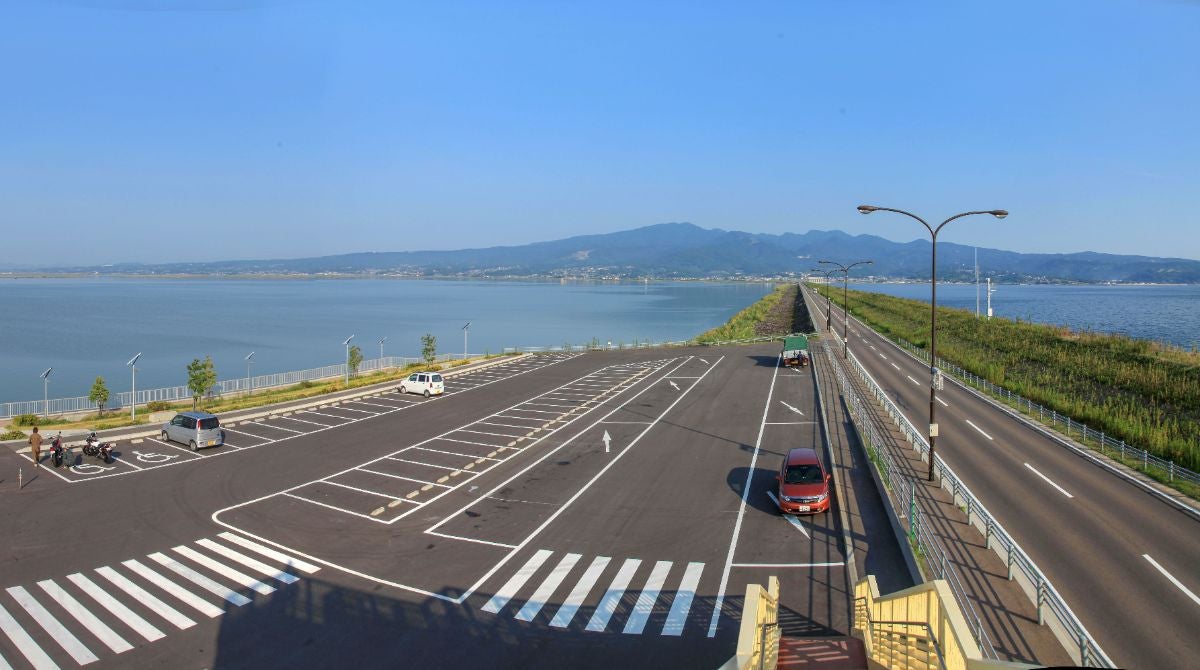 堤防道路の中間地点にある広い駐車スペース。展望歩道橋からの俯瞰。駐車場に数台のクルマが止まっており、その横を一直線のメイン道路が走っている。
