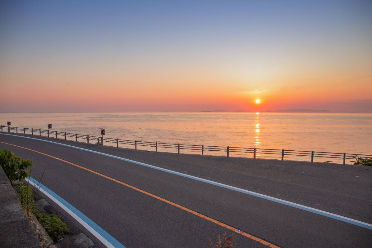 夕暮れ時、伊予灘に沈む太陽。空と海があかね色に染まり、水平線に夕日が沈んでいく。国道378号のガードレール越しに眺める、感動的な夕景。