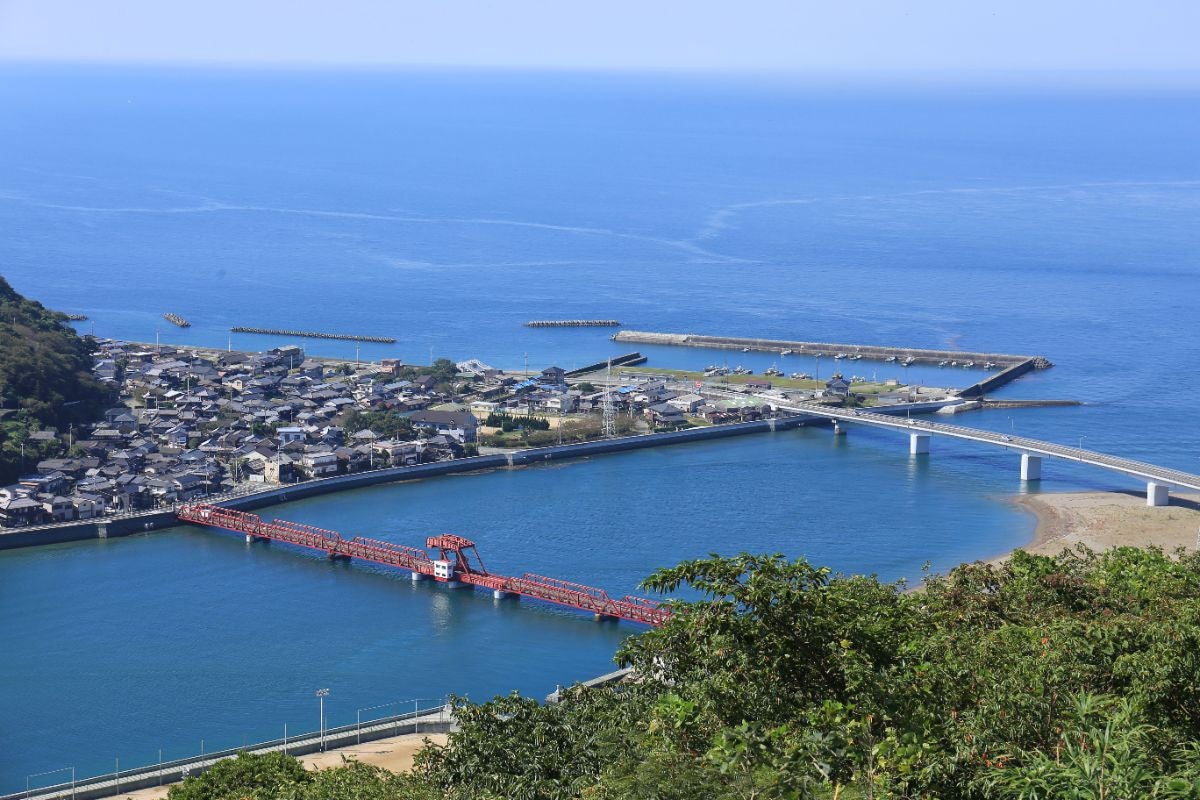 高台から望む、長浜大橋と伊予灘の青い海。赤いトラス構造の橋が川を横切り、対岸には長浜の町並みと港が広がる。遠くには水平線が続き、空と海が一体となったような爽快な景色であるコントラストが美しい。