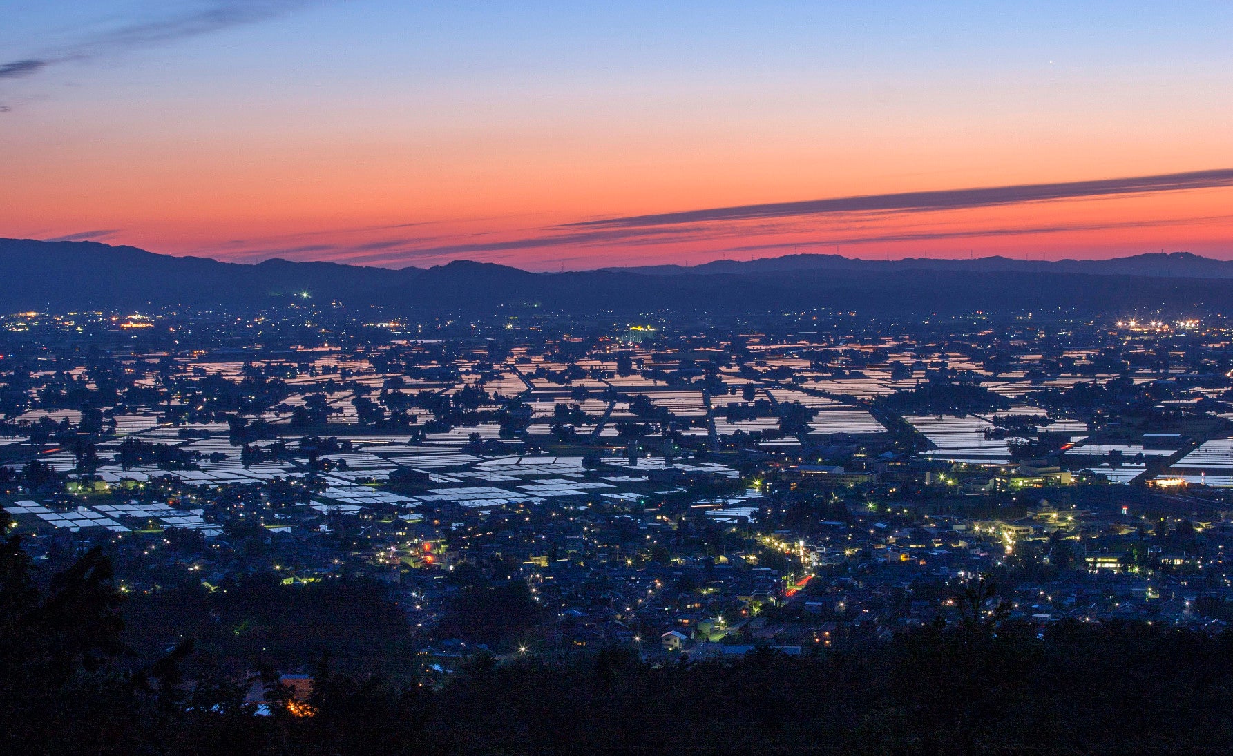 ALT 水が張られた田んぼが一枚一枚鏡のようになり、夕日のオレンジ色や空のグラデーションを反射する。この「水鏡」が広がる散居村の景色は、この場所ならではだ