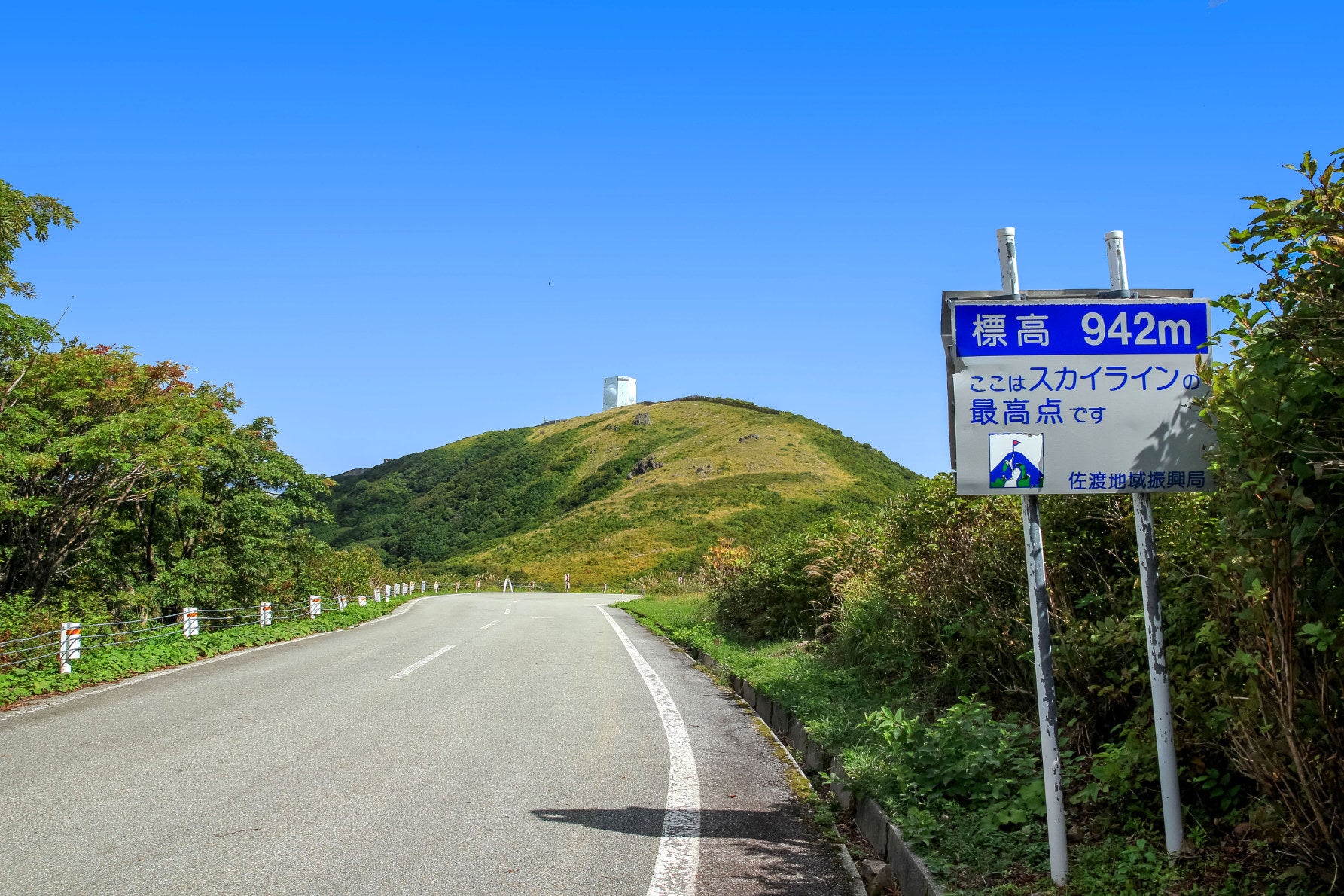 ALT 青空に向かって延びる大佐渡スカイラインの最高地点（標高942m）を示す案内看板。遠くの山頂付近には、特徴的な白い自衛隊のレーダーが見える