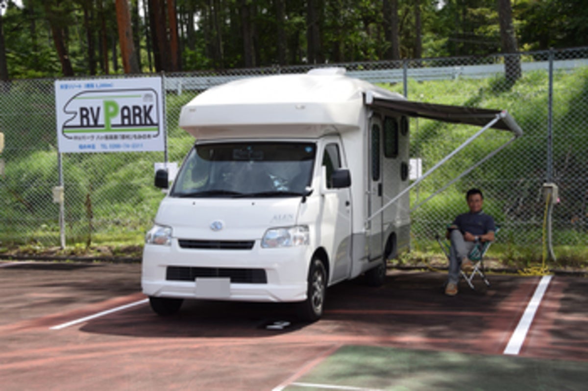 RVパークの区画内に停まったキャンピングカーと、サイドオーニングの下で椅子に座りくつろぐ男性の様子