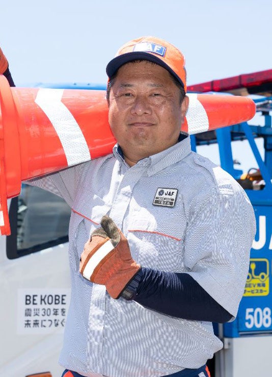 AF全国ロードサービス競技大会2025 関西本部代表・下園正和隊員（47歳・兵庫支部所属）｜パイロンを手に微笑む港隊員の姿と阪神大震災30年のステッカーを付けたレッカー車