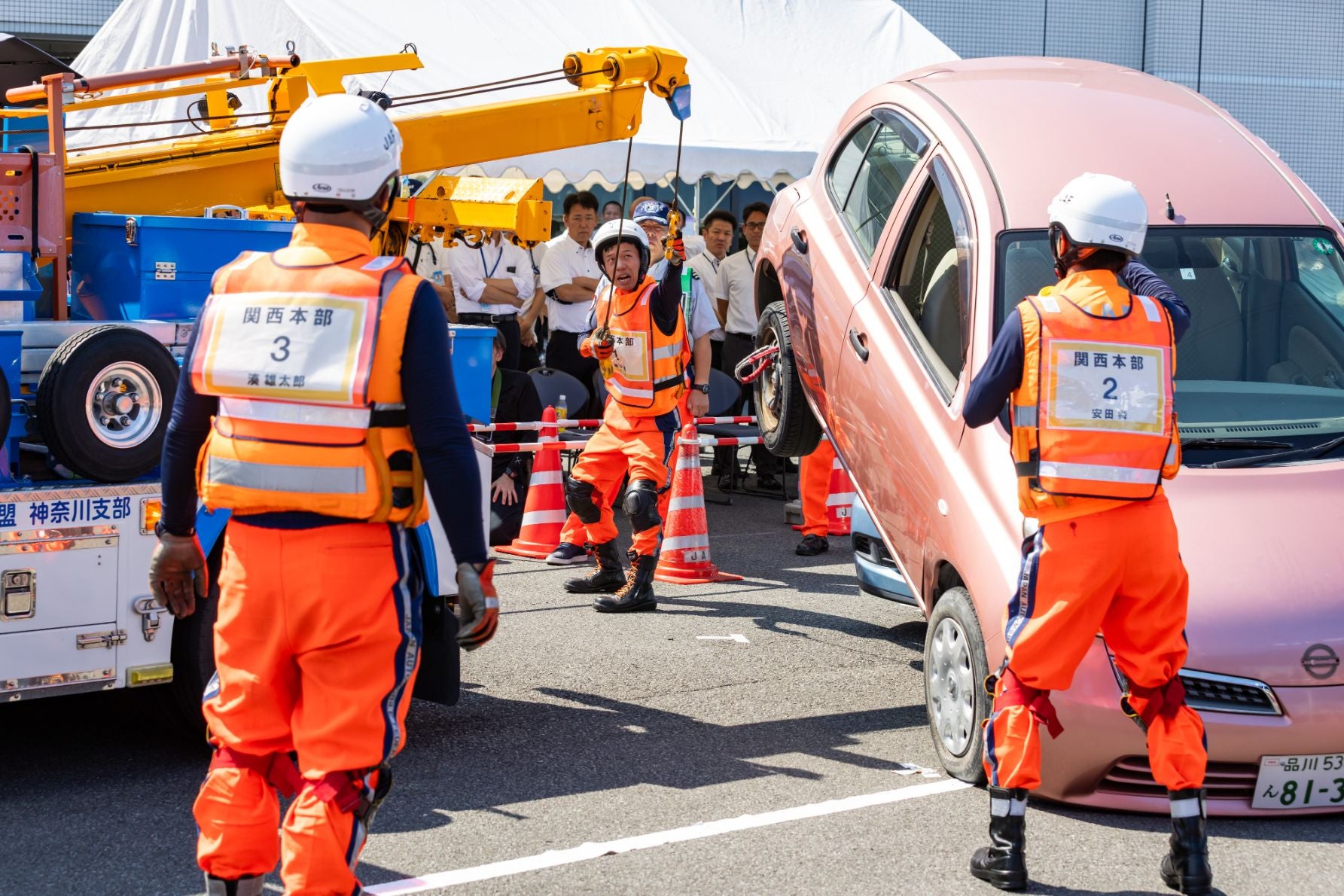 団体競技で、JAFのオレンジ色の制服を着た関西本部の選手がクレーンでピンク色の車両を持ち上げている。背景には白いテントと見物人が見える
