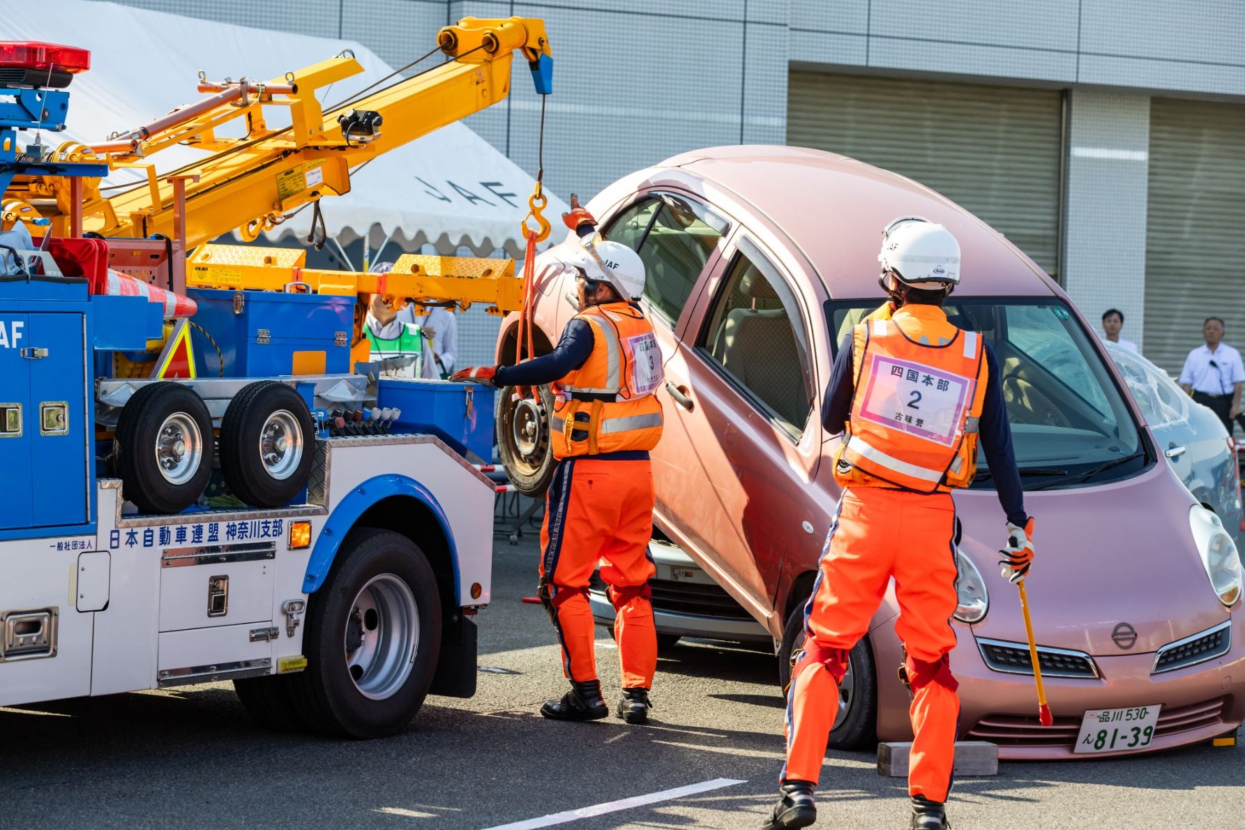 JAFのレッカー車がピンク色の車をけん引している屋外の様子。オレンジ色の制服とヘルメットを着用した指揮官の指示で作業員が操作を行い、周囲には見学者がいる