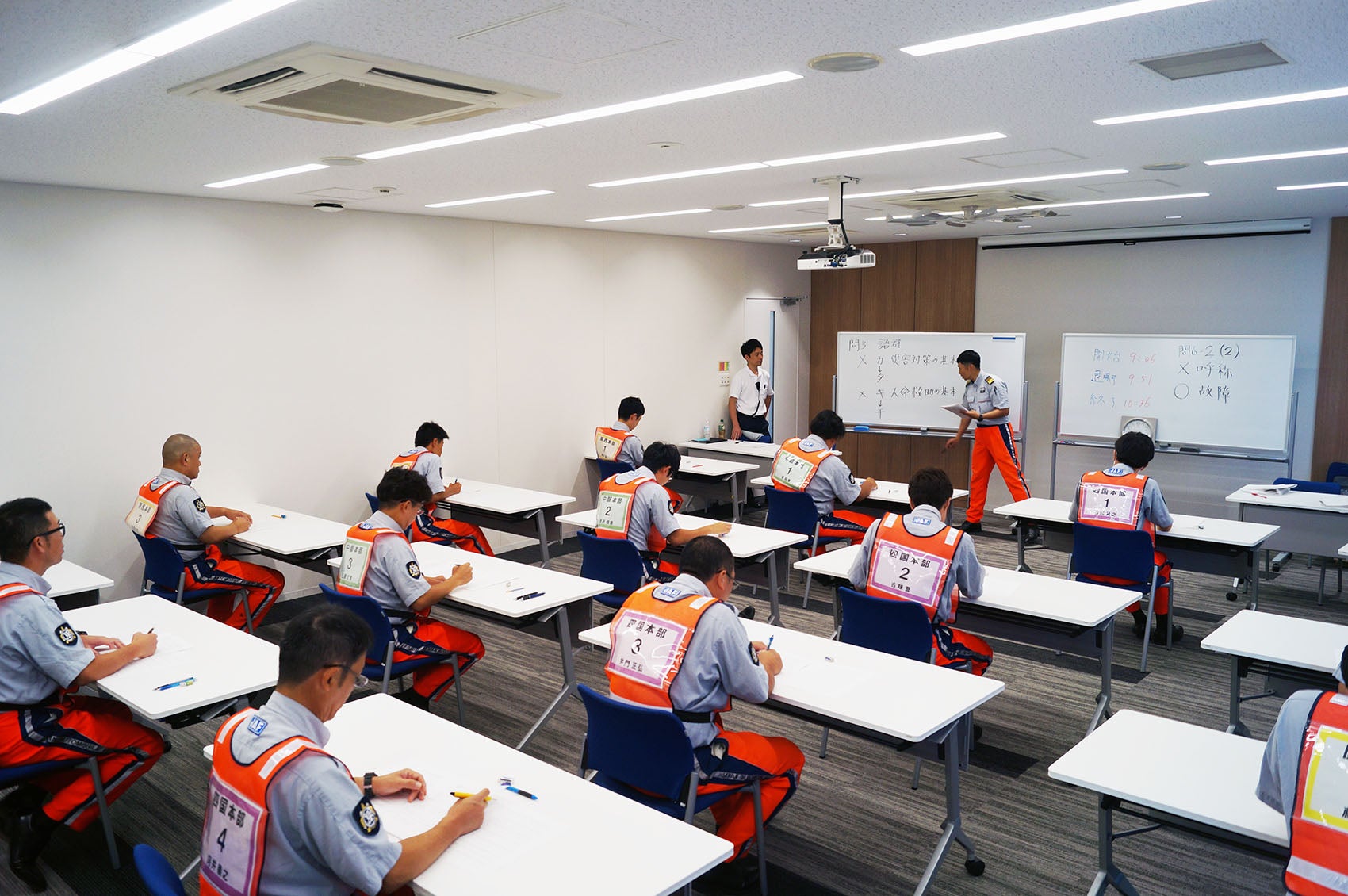 教室内でオレンジ色のJAFの制服を着た隊員が机に座り、試験を受けている。前方にはホワイトボードと講師が立っている