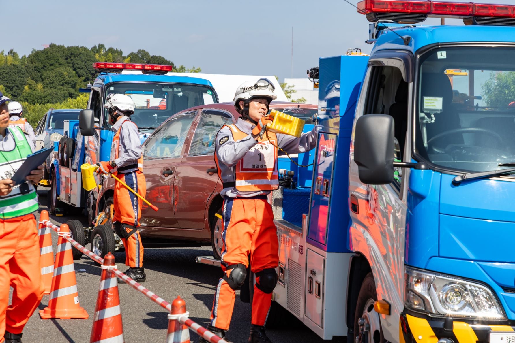 ヘルメットとオレンジ色の制服を着用した選手が、青いレッカー車の周囲で対応している。メガホンやクリップボードを持つ指揮官もおり、三角コーンで現場が囲まれている