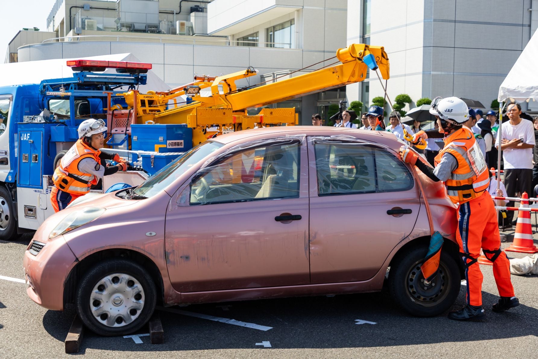 オレンジ色の制服を着たJAFの隊員が、レッカー車でピンク色の車をけん引する団体競技の様子。背景には見学者と三角コーンが見える。
