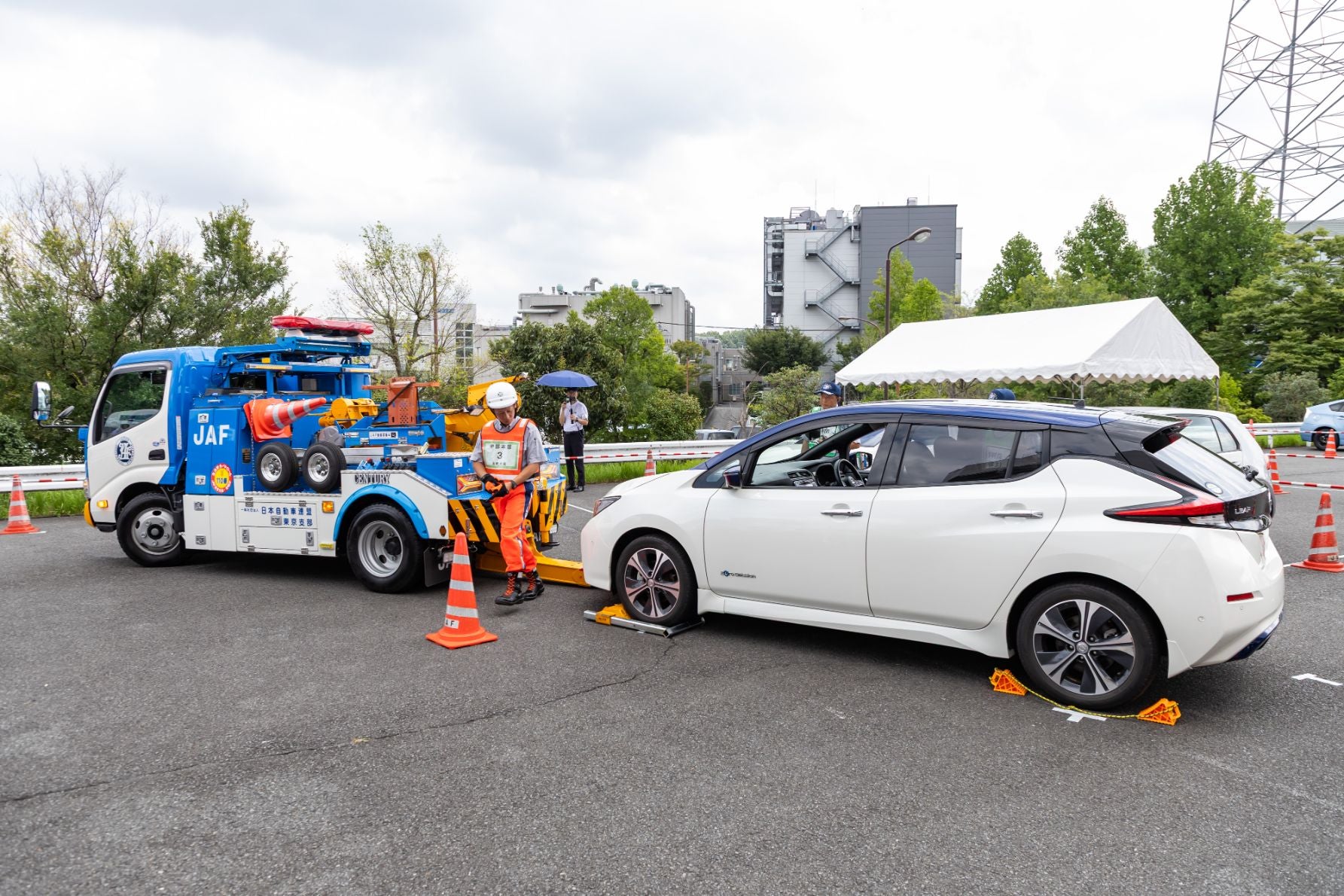 JAFのレッカー車が白い乗用車をけん引している様子。車のタイヤには輪止めが設置されており、周囲にはオレンジ色の三角コーンが配置されている。安全装備とヘルメットを着用した人物が車のそばに立っており、背景にはテント、木々、建物が見える。
