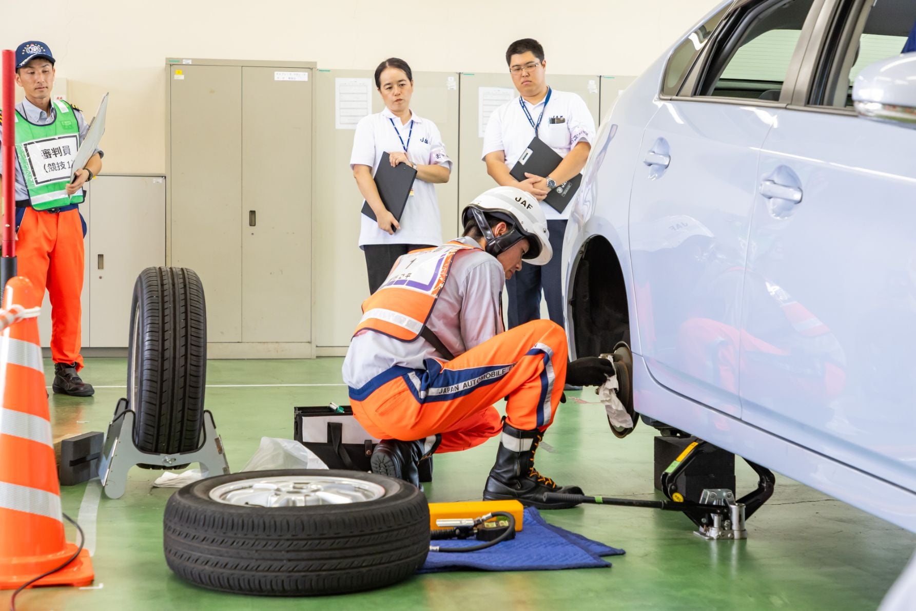 室内で安全装備を着用したJAFのロードサービス隊員が車のタイヤ交換作業を行っている。隊員は車の外れたホイールの横で膝をつき、工具を使って作業中。近くにはスペアタイヤと各種工具が置かれている。3人の作業を見守っており、背景には収納ロッカーと安全用の三角コーンが見える。
