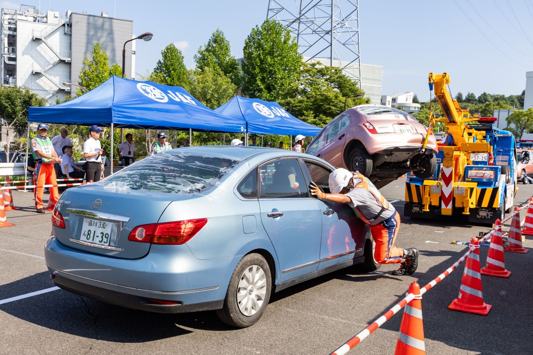 屋外の団体競技の現場で、青い車を作業員が押しており、背景ではJAFのレッカー車がピンクの車をけん引している。周囲には三角コーンやテープが設置され、JAFのテントの下に複数の人が作業を見守っている