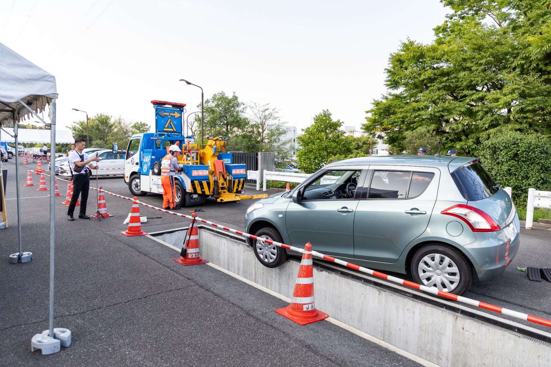 JAFのレッカー車のそばで隊員が車両けん引作業を行っている屋外の様子。周囲には三角コーンとバリケードが設置されている