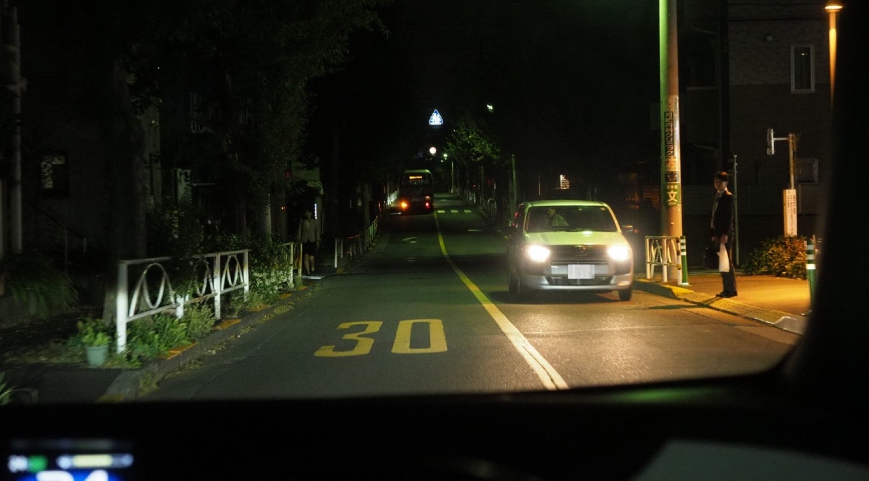 夜間、信号機のない交差点の手前で対向車とすれ違う自車。左右の歩道には歩行者がいる